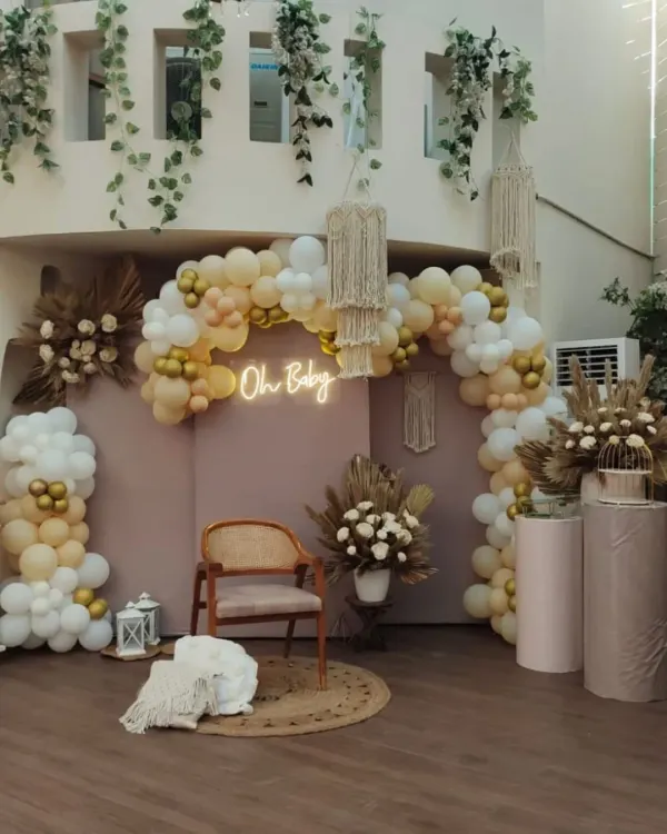 A complete boho-chic baby shower setup with an "Oh Baby" neon sign. The decor features a dusty pink backdrop, a balloon arch in neutral tones, macrame chandeliers, and a cozy seating area with a jute rug and woven throw.