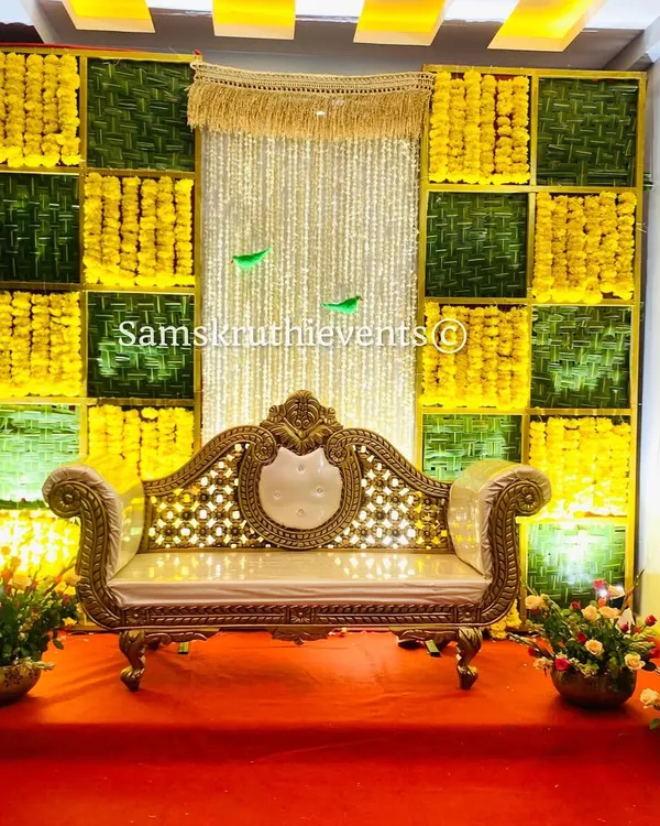 A classic Haldi or Mehendi backdrop featuring a checkerboard pattern of yellow marigolds and green woven leaves. The ornate gold and white sofa provides a perfect seat for the ceremony.
