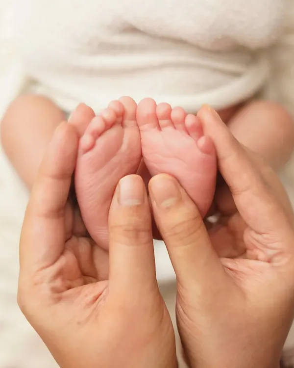 A close-up of a parent's hands gently holding their newborn's tiny feet. The image is shot from above against a soft white blanket, emphasizing how small and perfect they are.