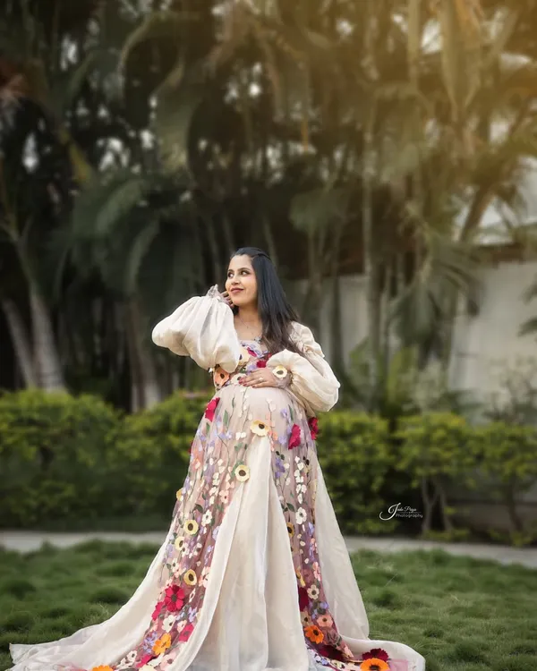 An outdoor maternity portrait glowing in the golden hour light. The expecting mother looks radiant in a beautiful floral embroidered gown, surrounded by lush greenery.