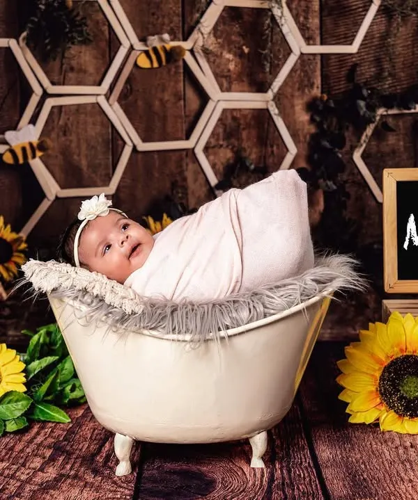 A bright and cheerful honeybee theme for baby Mehar. The newborn is awake and looking at the camera from a miniature tub, set against a honeycomb backdrop with sunflowers and a personalized nameplate.