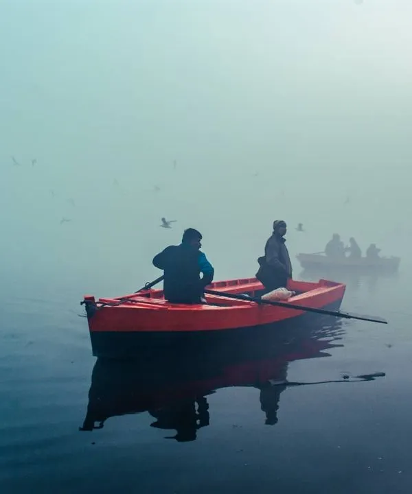 The bright red boat provides a striking pop of color against the foggy, muted tones of the Yamuna river. This contrast is what makes the scene so visually interesting.