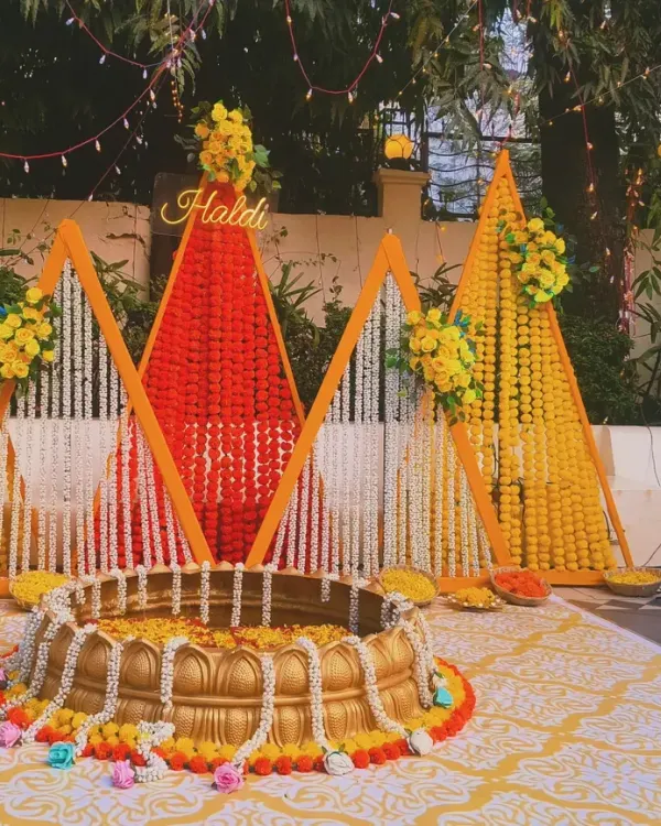 An outdoor Haldi ceremony setup with a backdrop of marigold strings in geometric patterns. The traditional brass urli in the center is filled with flowers for the ritual.