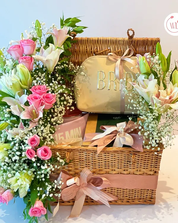Elevate the bride's experience with this stunning essentials package. Housed in a classic wicker picnic basket, it's filled with curated gifts and adorned with an explosion of fresh pink roses and lilies.