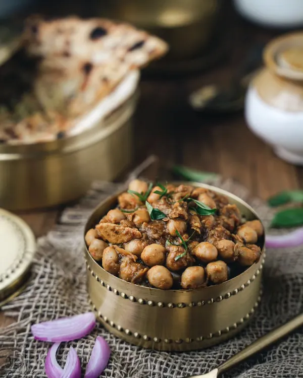 A moody, rustic shot of chole served in a traditional brass bowl. The dark lighting and textured surface bring out the rich colors of the curry, making it feel hearty and authentic.