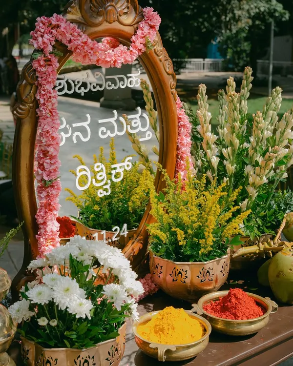 A personalized welcome mirror with the couple's names in Kannada script, surrounded by pink flowers and traditional elements like turmeric and kumkum.