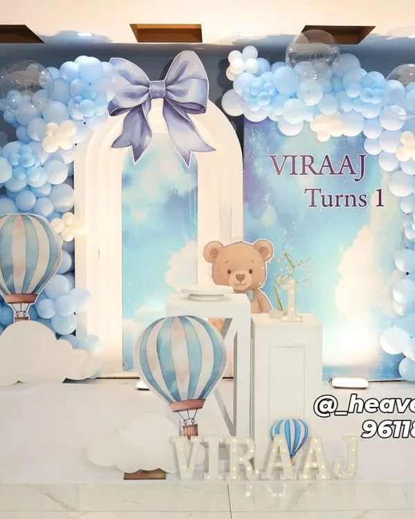 A dreamy hot air balloon and teddy bear theme for a first birthday, featuring a custom arched backdrop against a soft blue wall. The setup includes a full organic balloon arch in shades of blue and white, themed plinths, and large illuminated letters spelling out the name.