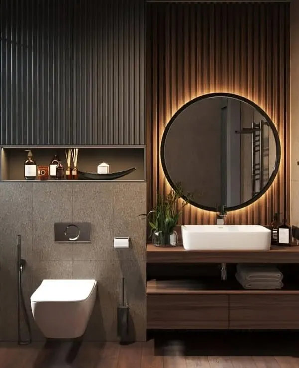 A contemporary bathroom featuring a custom floating wood vanity, a large backlit round mirror, and a mix of dark fluted and wood paneling on the walls.