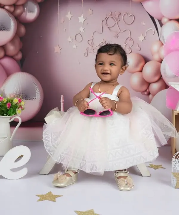 A playful shot from a first birthday milestone session. The birthday girl, dressed in a lovely white frock, shows off her personality with a pair of fun sunglasses, adding a touch of whimsy to her portraits.