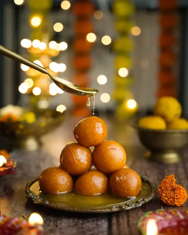 Happy Diwali. A classic festive shot of gulab jamuns stacked in a pyramid, with a spoon pouring syrup over them. The diyas and flower garlands in the background complete the scene.
