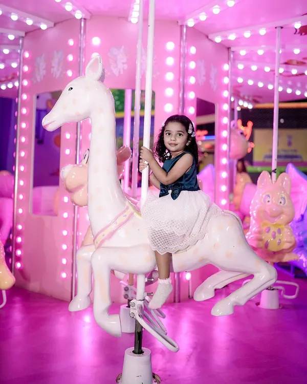 A magical moment from a child's birthday celebration, featuring a little girl riding a carousel horse. The bright pink lights of the merry-go-round create a whimsical and joyful atmosphere.