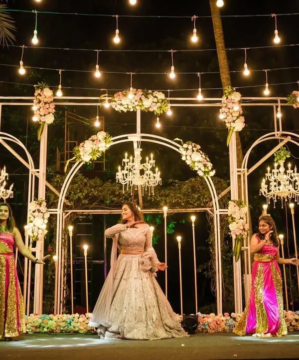 A spectacular stage for a Sangeet night performance. The design features white arches, multiple crystal chandeliers, and dramatic lighting to make every performance look like a professional show.