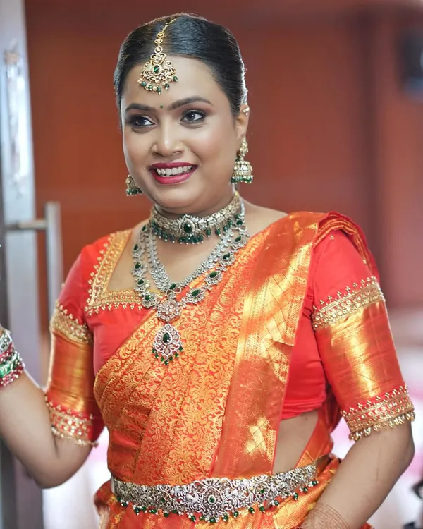 A radiant and happy bride at her engagement. This look features glowing skin, defined eyes, and a bright smile, perfectly matching her vibrant orange Kanjivaram saree and layered diamond jewellery.
