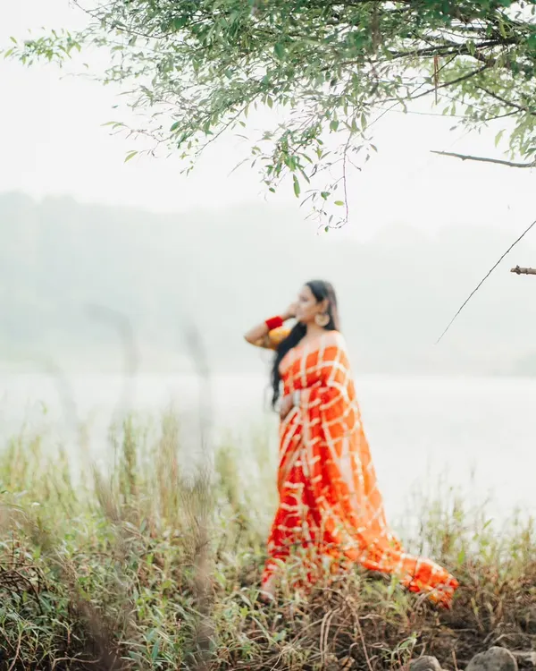 An artistic, soft-focus shot of a woman in a vibrant orange bandhani saree by a lake. This image has a dreamy, ethereal quality.
