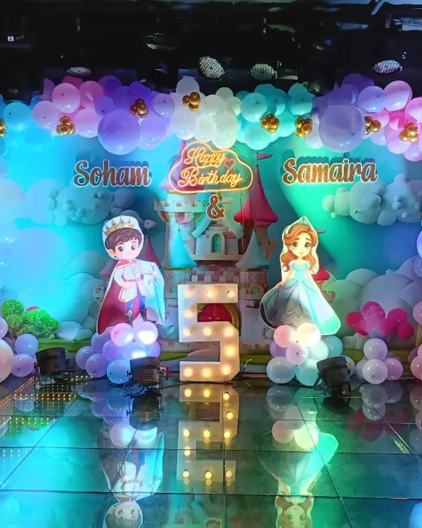 A full stage setup for a royal 5th birthday party. This Disney Princess theme features character cutouts, a castle backdrop, and a massive balloon arrangement in pastel colors, all illuminated with stage lighting for a dramatic effect.