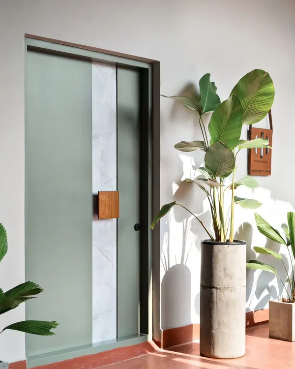 The entrance to this office was designed to be a serene transition space. The soothing green door features a stone laminate inlay and a solid teakwood handle, creating a simple yet elegant statement. The surrounding plants and soft gray wall add to the calm, welcoming ambiance.