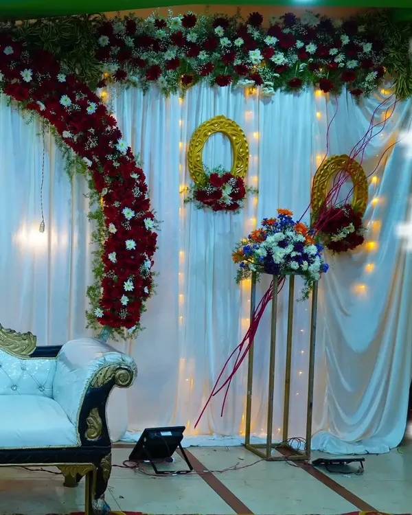 A classic and elegant stage design for a wedding or reception. This setup features a rich red and white floral arch, matching floral arrangements, and a backdrop of white curtains with fairy lights.