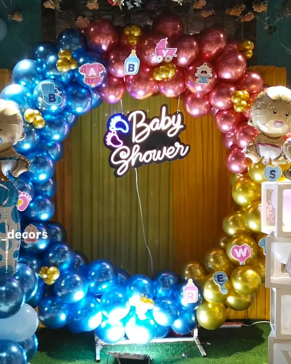 A full circular balloon arch in blue, pink, and gold creates a stunning photo booth for a baby shower. The glowing 'Baby Shower' neon sign in the center adds a modern and festive touch to the celebration.