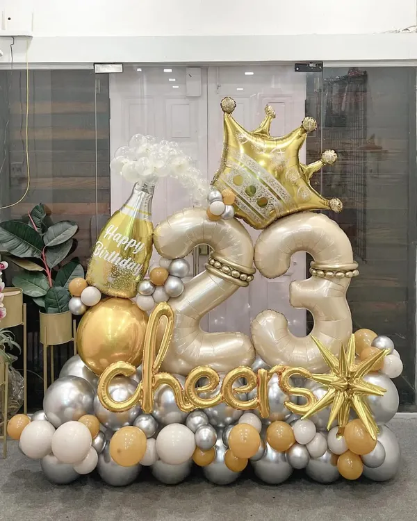 Celebrate turning 23 in style with this luxe arrangement. Featuring a popping champagne bottle, a golden crown, and "cheers" script, this display in a sophisticated silver and gold palette is designed to feel like a true royal celebration.