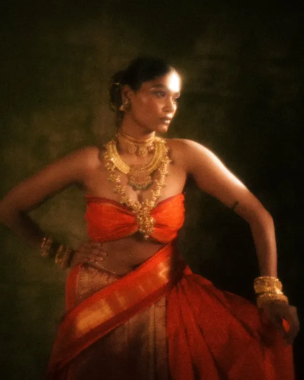 A powerful stance from the 'Swaṟṇavalli' series. The warm, hazy lighting beautifully complements the gold jewelry and the rich orange of the sari, while the makeup remains sharp and defined.