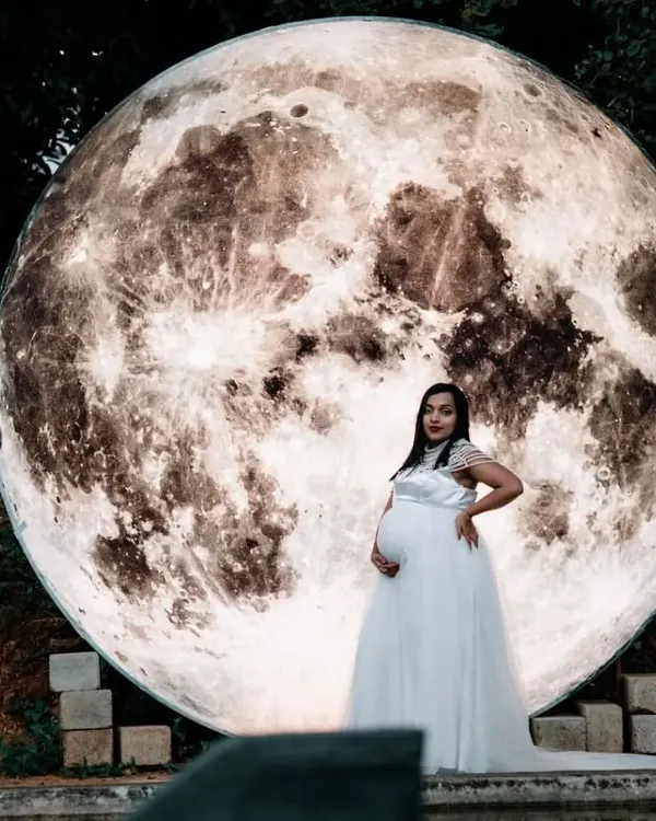 A breathtaking solo portrait of a mother-to-be in a white gown, standing before our giant illuminated moon prop. This is one of our signature creative concepts for an unforgettable night shoot.