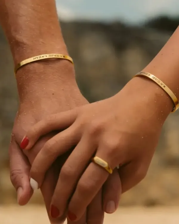 A couple holding hands, both wearing matching gold-plated cuff bracelets engraved with special coordinates. This is a beautiful way to commemorate a shared place or memory.