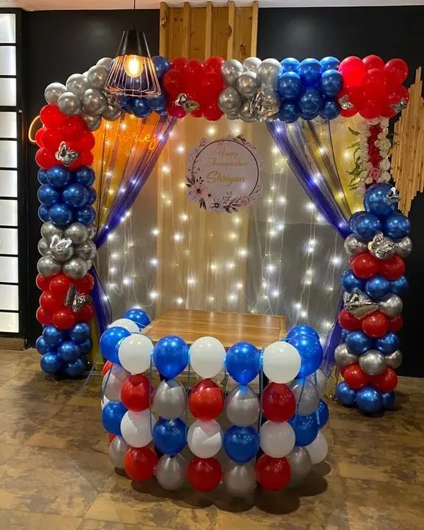 A bold Annaprashan ceremony decoration with a balloon pillar arch in red, blue, and silver. The setup includes a backdrop with fairy lights and a custom sign for the ceremony.
