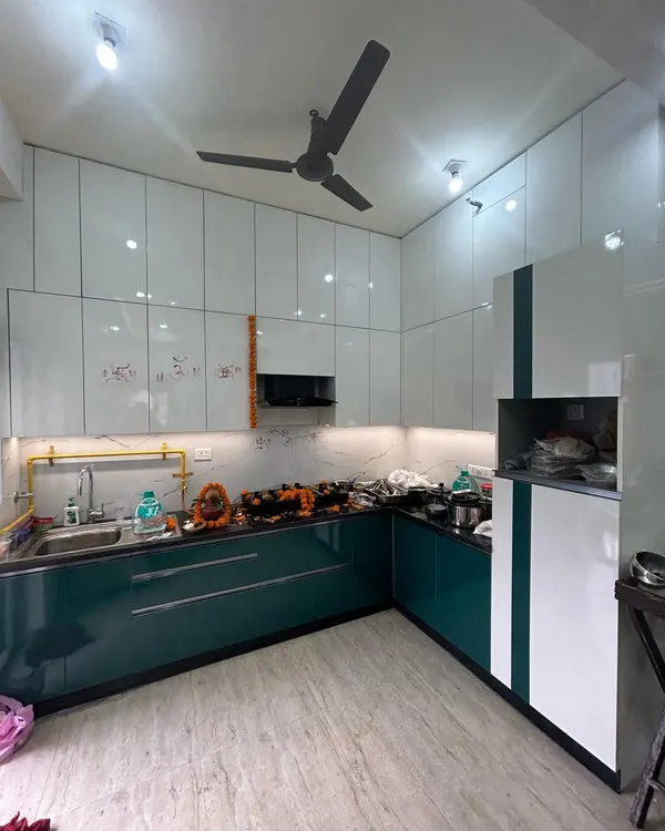 This recently completed 3BHK kitchen features a stunning combination of teal green and glossy white cabinets. The design includes a built-in chimney, under-cabinet lighting, and a marble-patterned backsplash for a clean, contemporary look.