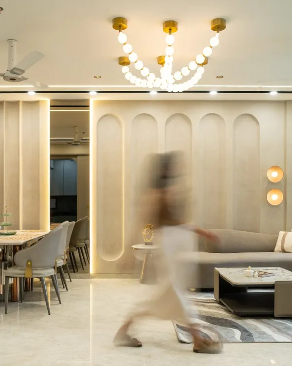 This luxurious living-dining space embraces fluidity through its curved wall paneling and layered lighting. A sculptural chandelier anchors the ceiling, while soft neutral tones, brushed gold accents, and plush textures create a seamless blend of modern sophistication and quiet opulence.
