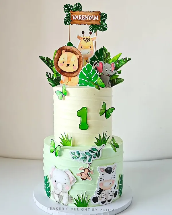 A dreamy two-tier jungle safari cake in soft pastel tones for a "Wild One" first birthday. This design uses a mix of 2D and 3D elements, including baby lions, giraffes, and elephants, to create a gentle and charming look.
