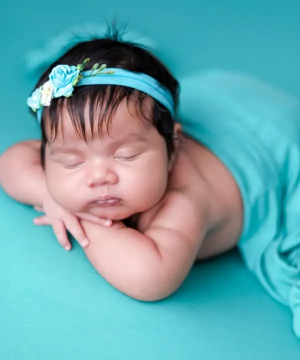 The 'chin on hands' pose is a newborn photography classic. We achieve this pose safely while the baby is in a deep sleep, using a vibrant teal backdrop and matching headband.