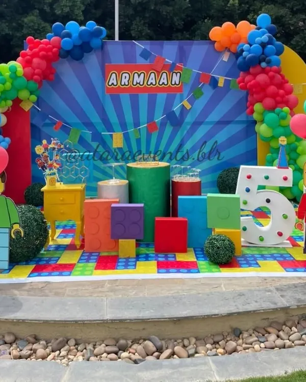 Welcome to a world of colorful bricks! This full stage setup for a 5th birthday features giant LEGO blocks, character cutouts, a light-up number, and a vibrant balloon arch.