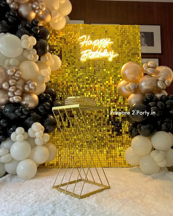 A sophisticated birthday setup featuring a gold sequin wall and a neon sign. The look is completed with an elegant balloon garland in black, white, and champagne gold, and a modern cake stand.