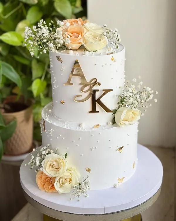 A full view of a stunning two-tier engagement cake. This design combines soft peach and white roses, baby's breath, and a custom gold 'A&K' monogram for a truly personalized statement.