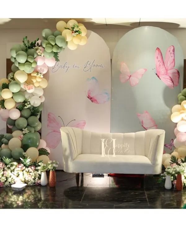 A 'Baby in Bloom' baby shower with a beautiful butterfly theme. The setup includes custom arched backdrops, a comfortable sofa for the mom-to-be, and an elegant organic balloon garland in sage green and pastel pink.
