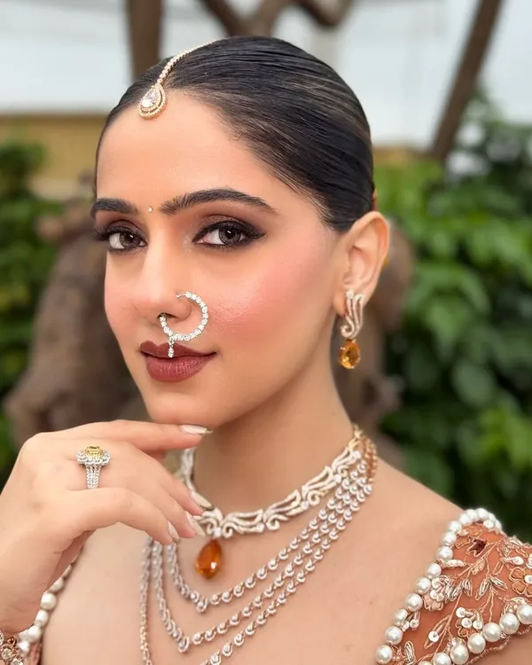 A close-up of this modern bridal look, focusing on sharp, defined eyes and a flawless skin finish. The diamond nath and layered jewelry perfectly complement the rust-orange lehenga, creating a look that is both contemporary and timeless.