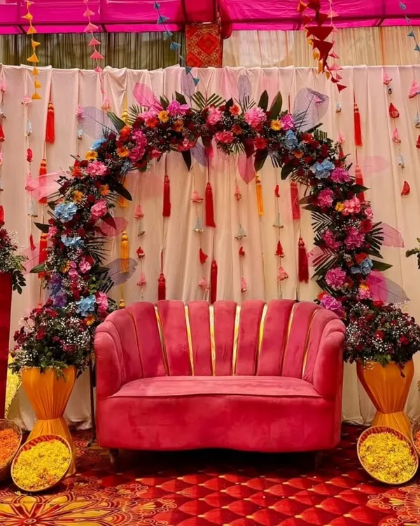 This vibrant Mehandi seating features a plush pink sofa set against a backdrop of colorful tassels and a dense floral arch. The setup includes baskets of fresh marigold petals for the rituals.