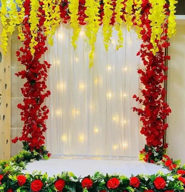 A grand arch-style backdrop made with bright red and yellow artificial flowers. The center features a sheer white curtain embedded with fairy lights, providing a luminous and festive background for your Ganpati idol.