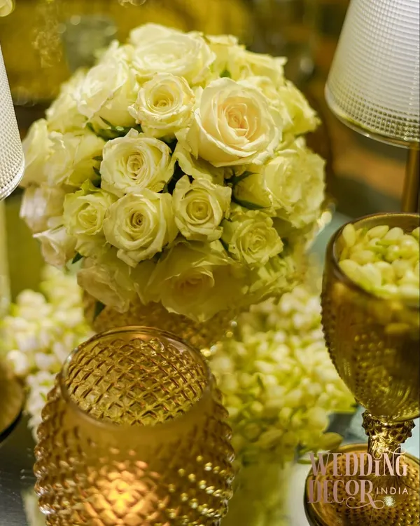 A close-up of a classic and elegant centerpiece. A tight bouquet of off-white roses is surrounded by the warm glow of candles in textured gold holders, creating a timeless and romantic look.