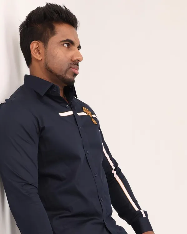 This navy blue shirt features a clean white stripe that runs across the chest and down the sleeve, accented by a small, intricate golden crest. It's a design that balances modern lines with classic, regal details.