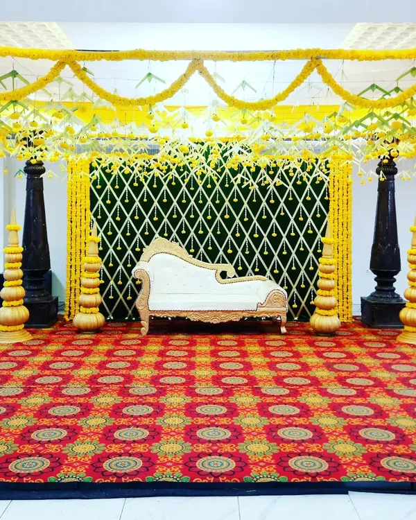A full view of a traditional mantap setup, perfect for pujas or wedding rituals. The design is defined by its dark, carved pillars, a canopy of yellow marigolds, and a vibrant patterned carpet.