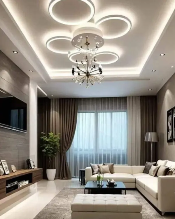 This living room features a complex and modern false ceiling design with a unique circular LED fixture combined with a chandelier. The multi-layered lighting creates a bright yet comfortable environment.