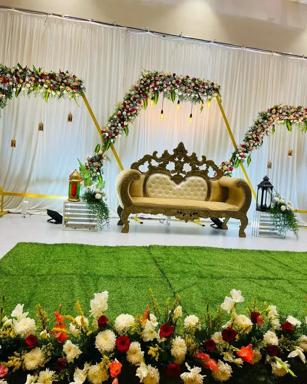 A modern and elegant stage setup for an engagement. The design features geometric arches decorated with flowers and hanging lights, with an ornate sofa as the centerpiece.