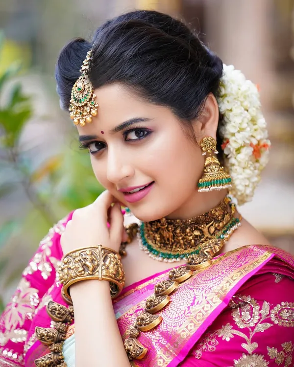 A classic South Indian bridal portrait of actress Ashika Ranganath, focusing on the rich colors of the saree and the exquisite detail of the antique gold jewelry.