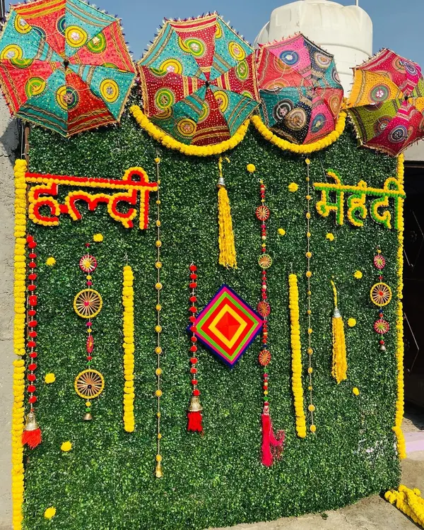 An artificial green wall provides a lush, modern base for this Haldi and Mehndi backdrop. I use real marigold flowers to write out "Haldi" and "Mehndi" and add traditional hangings and colorful Rajasthani umbrellas for a festive look.