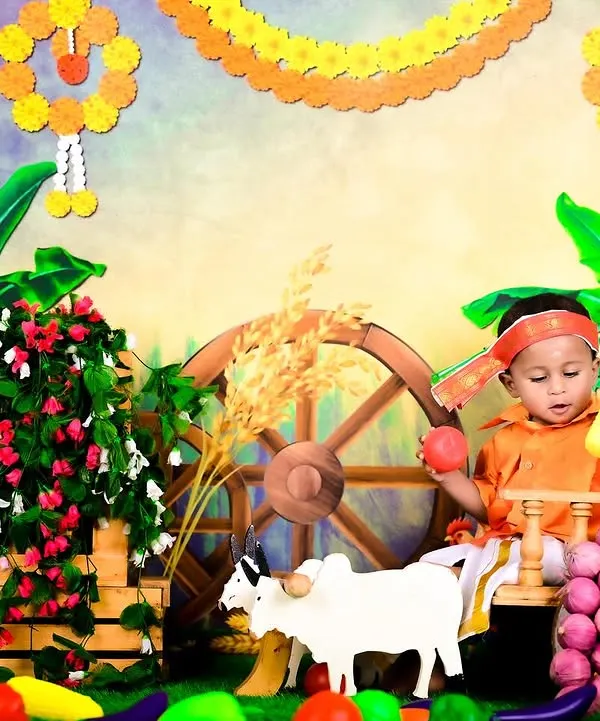 A baby dressed in traditional attire for a Sankranti or harvest festival theme, complete with a bullock cart and fresh produce.