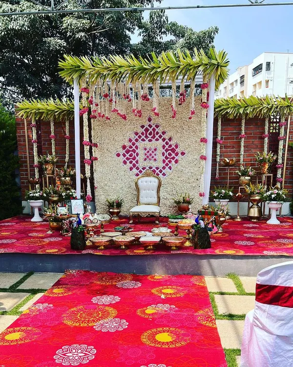 A beautiful outdoor stage setup for a Seemantham (traditional baby shower). The backdrop is a wall of white flowers with a pink floral pattern, set against a rustic brick wall.