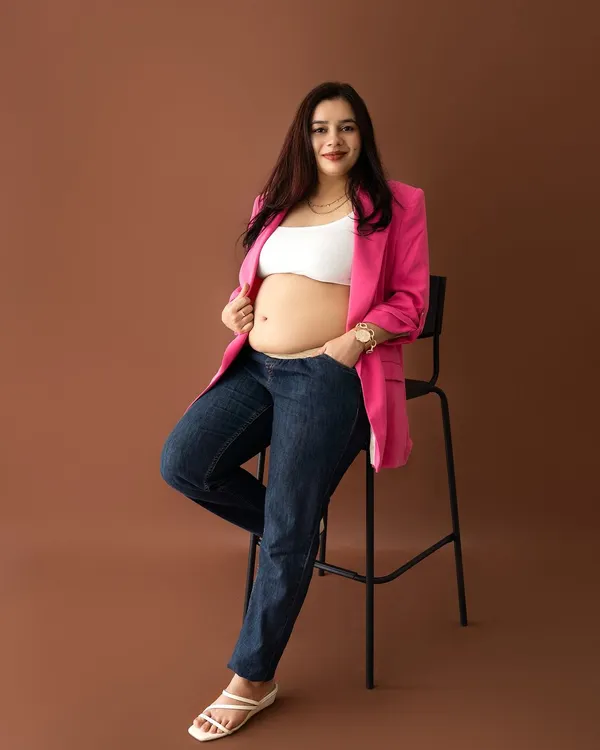 A confident and stylish maternity portrait of a mom-to-be in a pink blazer and jeans, leaning against a stool.
