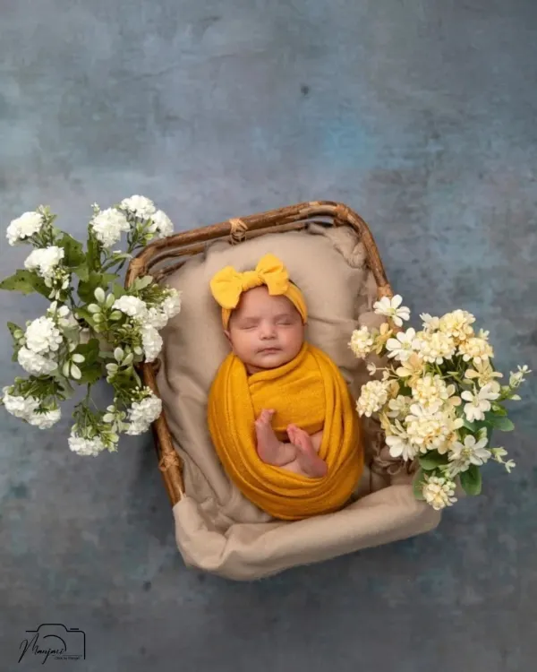 A vision in sunny yellow. This baby girl is nestled in a rustic basket, surrounded by delicate white and yellow flowers, creating a bright and cheerful newborn portrait.