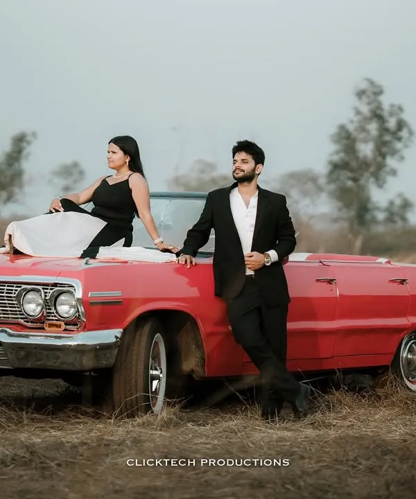 A stylish, wide-angle shot of a couple posing with a red vintage convertible in a field, capturing a classic, cinematic pre-wedding vibe.